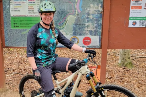 Amy on her bike posing in front of a bike trail in the mountains